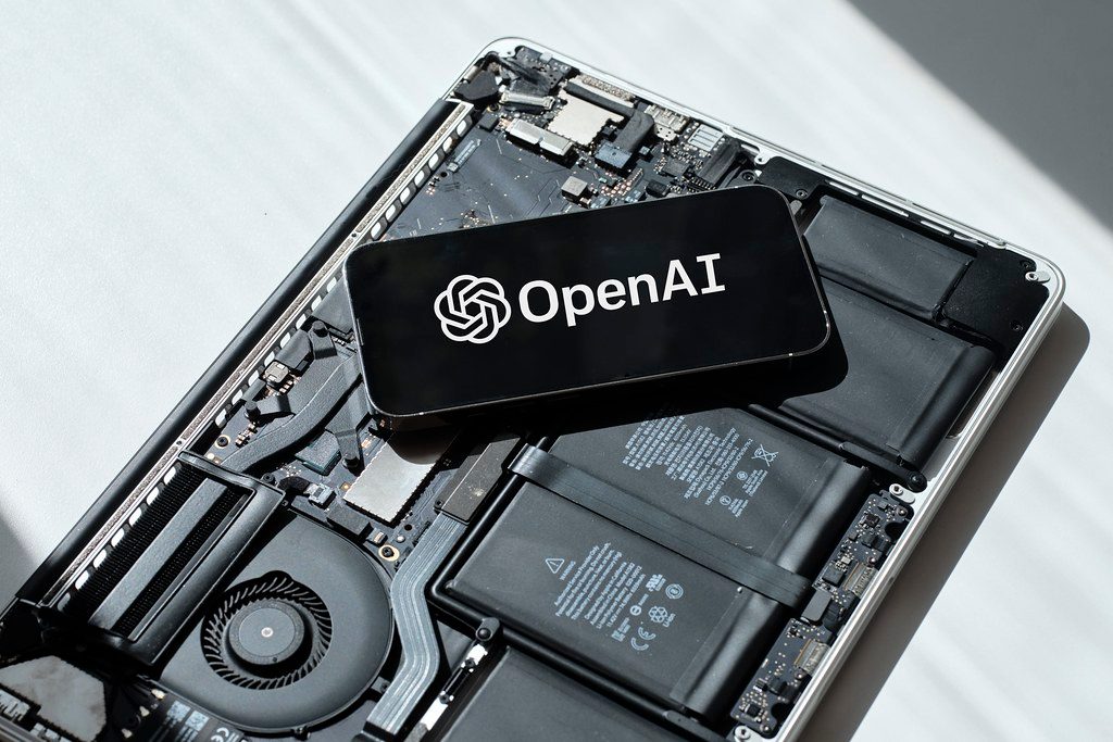 A smartphone displaying the OpenAI logo held in front of a blurred screen